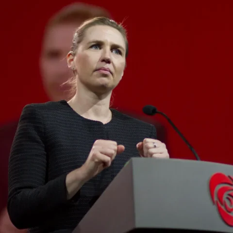 Foto de Mette Frederiksen em pronunciamento oficial, com fundo institucional. O texto aborda reforço militar da Dinamarca e OTAN no Ártico, em resposta à pressão dos EUA sobre a Groenlândia.